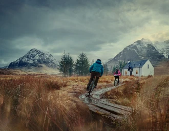 Two cyclists ride mountain bikes along a muddy path toward a white cottage beneath snow-capped mountains and a dramatic cloudy sky.