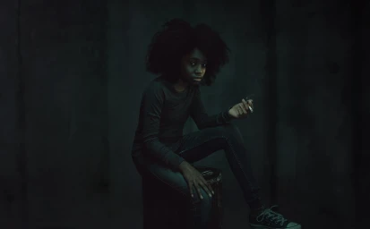 Black woman with voluminous dark hair seated on a stool holding a cigarette in a dark moody interior photographed by Todd Antony.