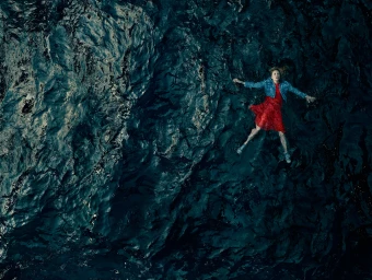 Aerial view of a person in a red dress and denim jacket floating on dark turbulent water.
