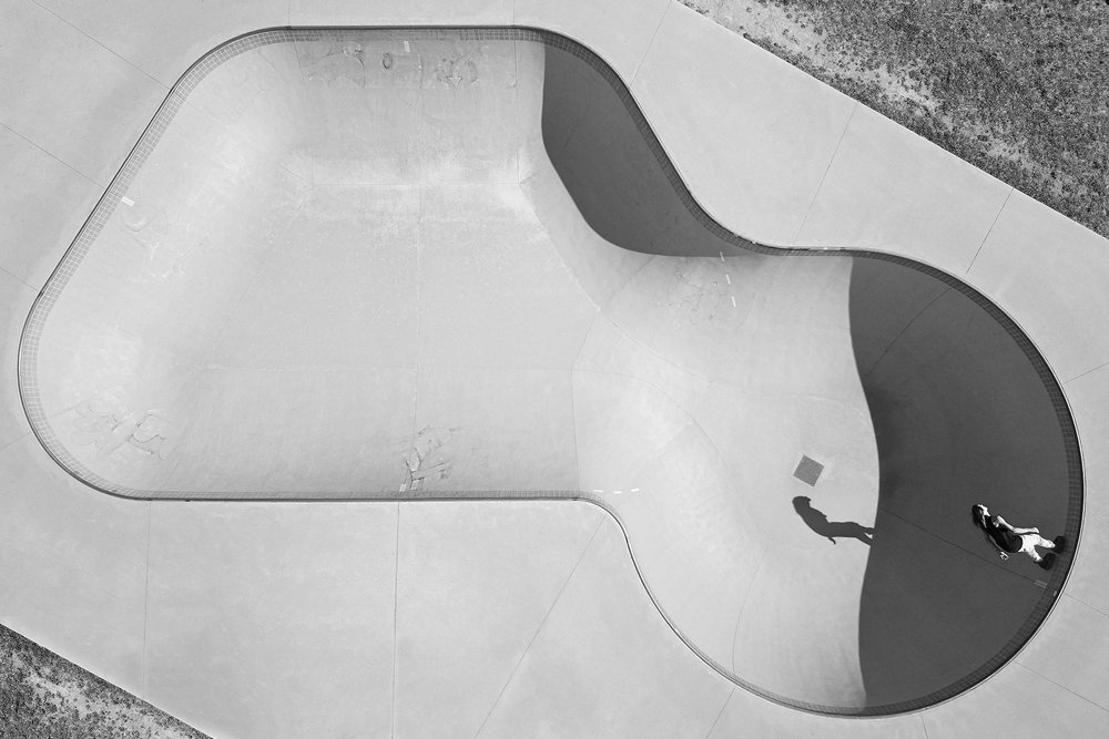 High-angle black and white photograph of a person skating in a concrete skatepark bowl with a square pattern on the concrete.