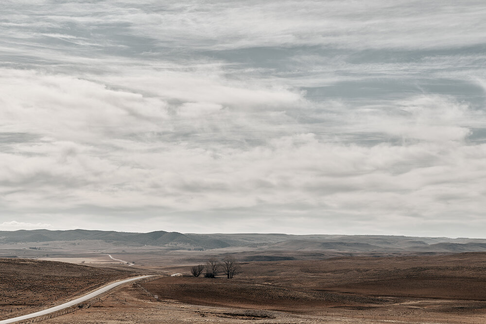 Panoramic landscape featuring a long road winding through brown fields under a vast, cloudy sky. A few trees are visible on the horizon.