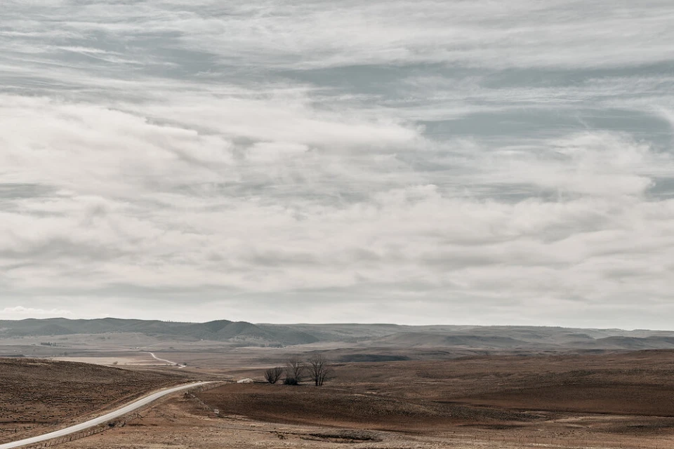 Panoramic landscape featuring a long road winding through brown fields under a vast, cloudy sky. A few trees are visible on the horizon.