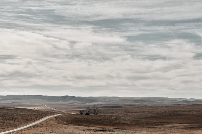 Panoramic landscape featuring a long road winding through brown fields under a vast, cloudy sky. A few trees are visible on the horizon.