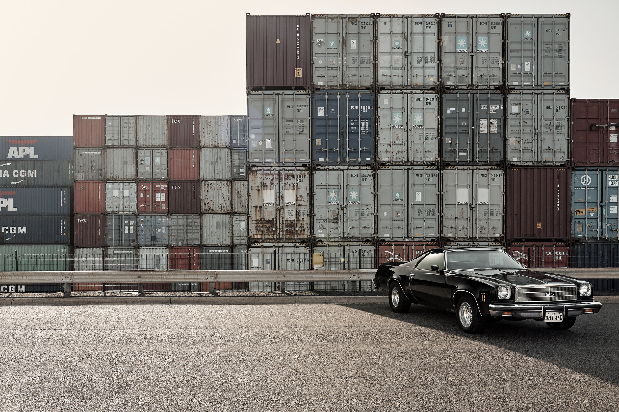 A vintage black coupe is parked on an asphalt surface against a backdrop of stacked shipping containers on a cloudy day.