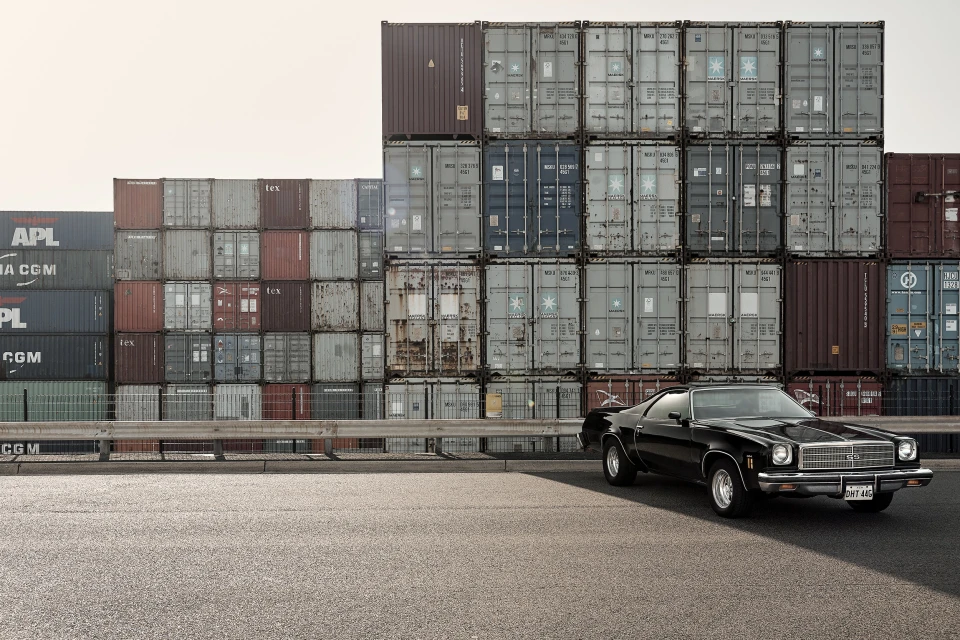A vintage black coupe is parked on an asphalt surface against a backdrop of stacked shipping containers on a cloudy day.