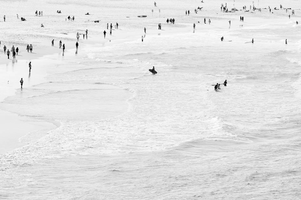 Monochrome beach scene with numerous people walking on the sand and wading in the ocean surf, captured from an elevated perspective.