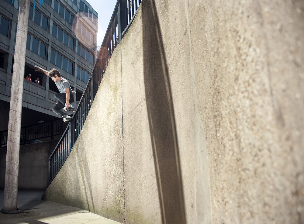 A skateboarder performs a trick mid-air near a concrete structure against a backdrop of an urban city building with window details.
