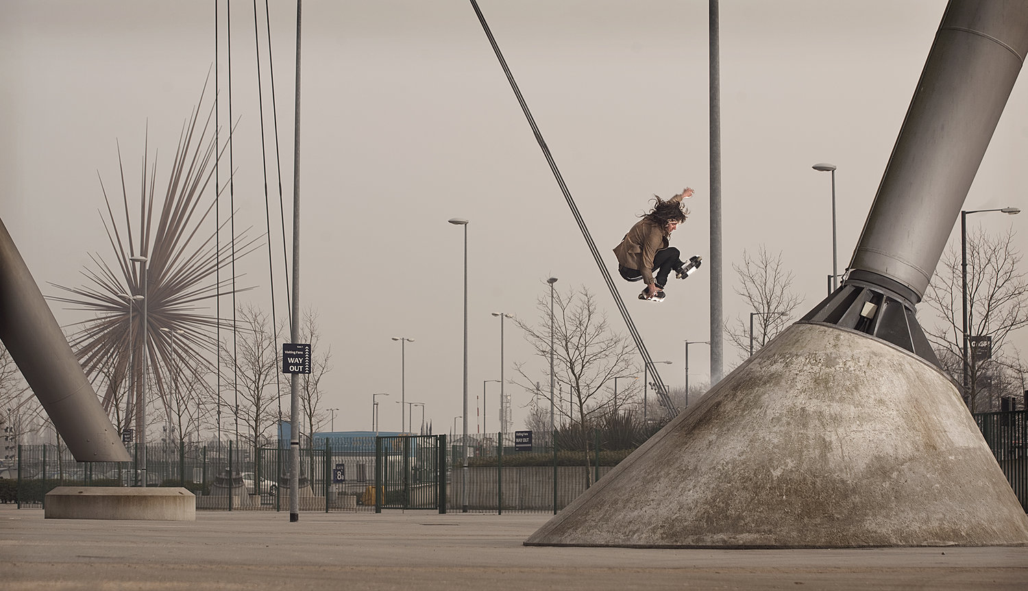 A skateboarder in mid-air performs a trick near a support structure; urban sports scene, overcast day, focus on action and architectural elements.
