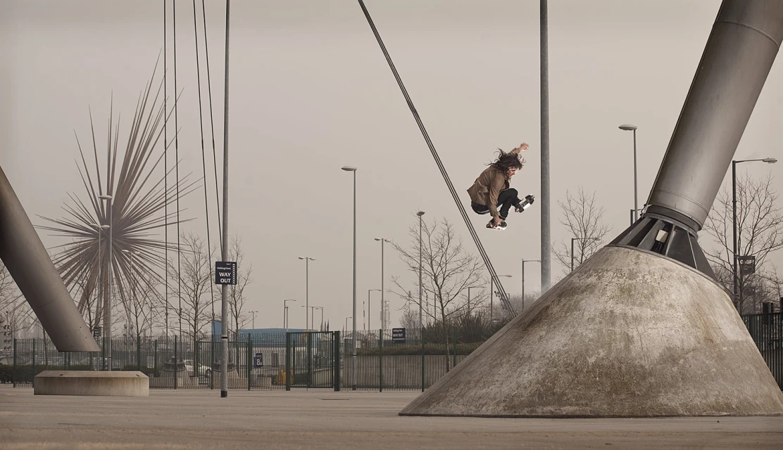 A skateboarder in mid-air performs a trick near a support structure; urban sports scene, overcast day, focus on action and architectural elements.