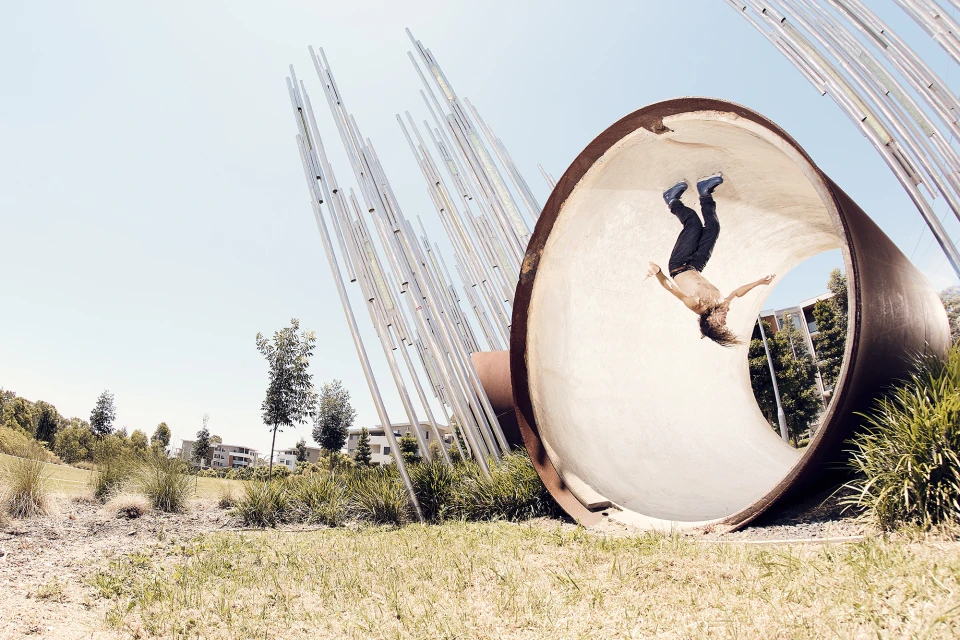 Adult male parkour athlete wearing roller blades performs a flip inside of a large concrete pipe in a grassy park on a sunny day.