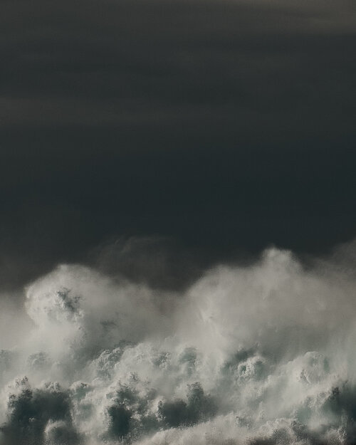A dynamic cloudscape shows turbulent white clouds against a dark grey sky, creating a dramatic natural scene with a sense of scale.
