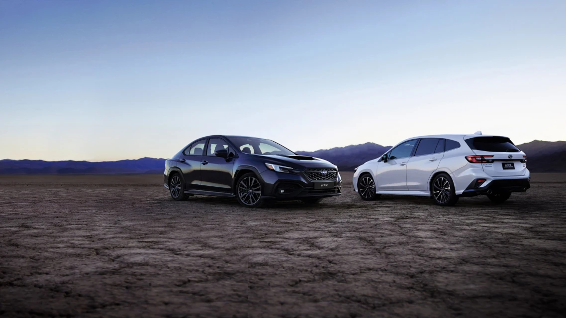 Two Subaru WRX cars, one black and one white, parked on a cracked desert surface with distant mountains under a pale blue sky.