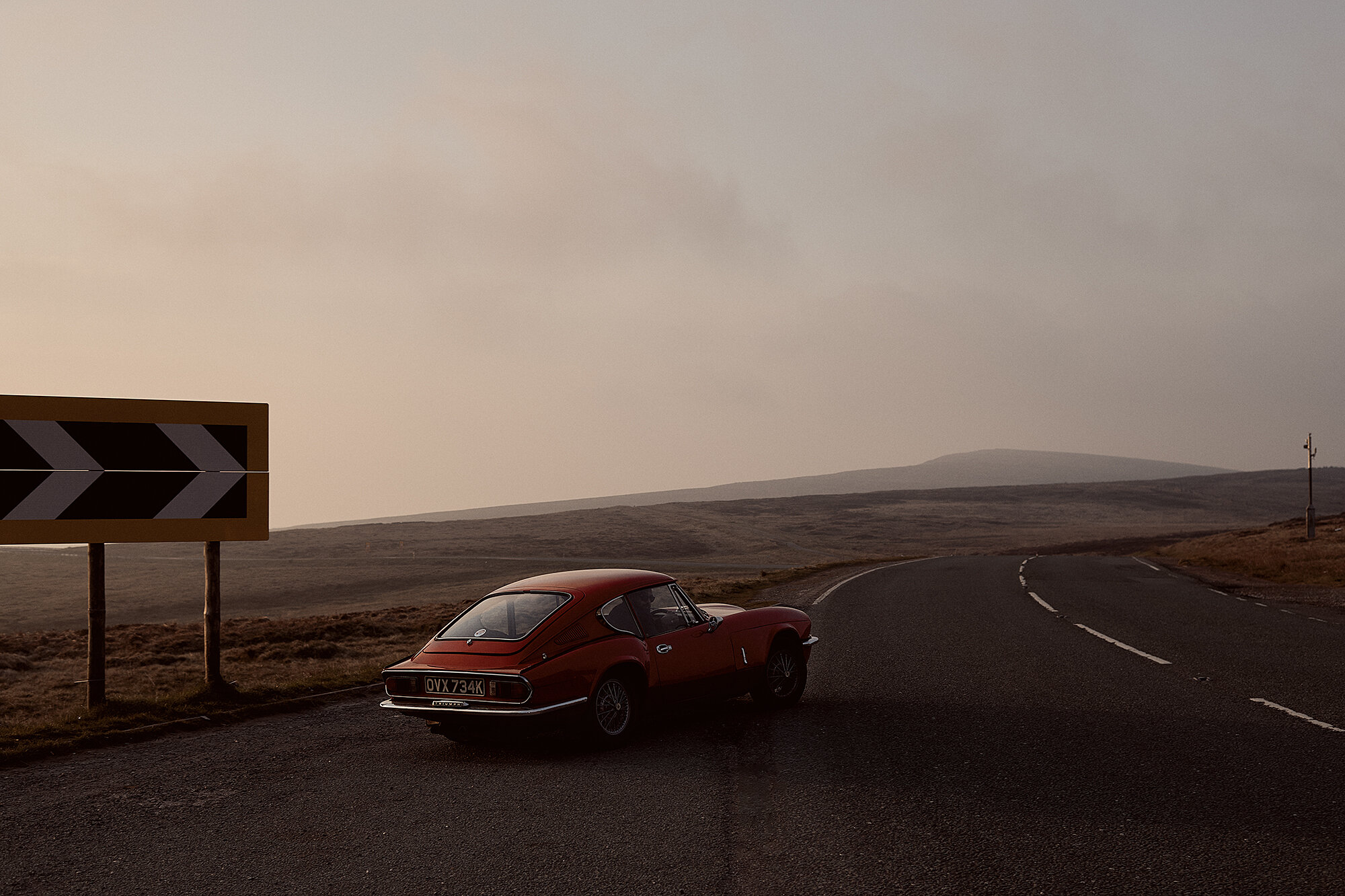 Red Triumph Spitfire sports car parked on an empty country road with a hairpin turn sign, set against a moody landscape and hazy sky.