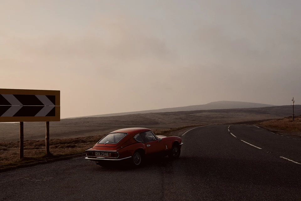 Red Triumph Spitfire sports car parked on an empty country road with a hairpin turn sign, set against a moody landscape and hazy sky.