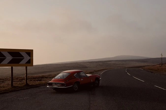 Red Triumph Spitfire sports car parked on an empty country road with a hairpin turn sign, set against a moody landscape and hazy sky.