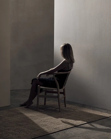 Woman with blonde hair seated on a wooden chair in a sunlit interior space with a textured rug and concrete walls