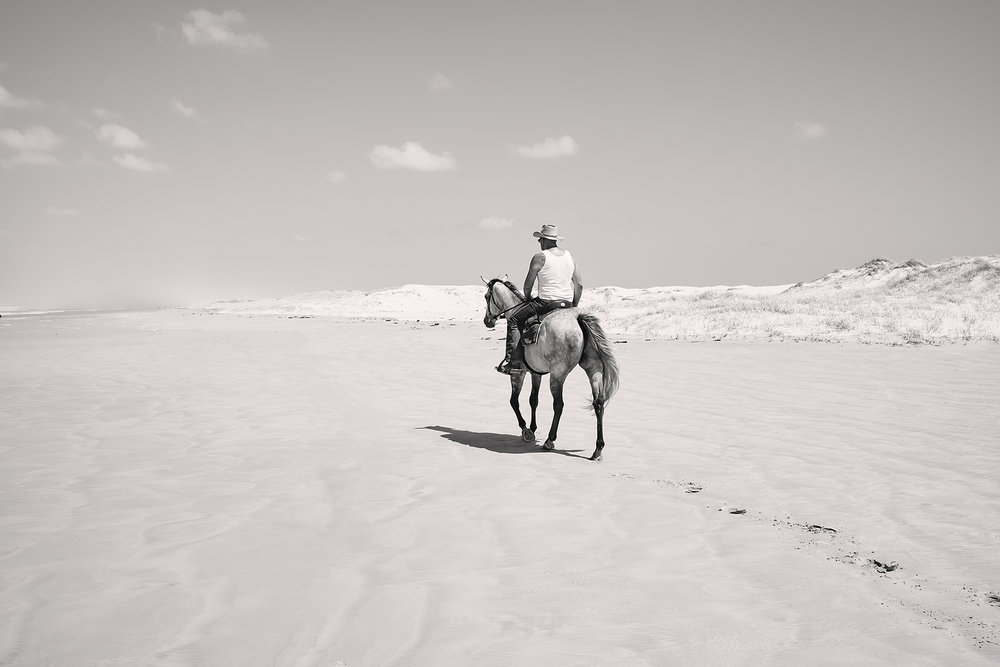 Monochrome image of an adult wearing a cowboy hat riding a horse along a sandy beach with dunes in the background and a bright, clear sky.