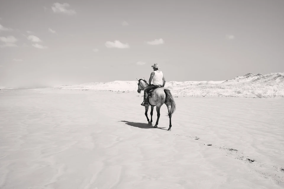 Monochrome image of an adult wearing a cowboy hat riding a horse along a sandy beach with dunes in the background and a bright, clear sky.