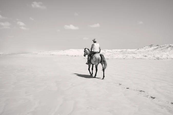 Monochrome image of an adult wearing a cowboy hat riding a horse along a sandy beach with dunes in the background and a bright, clear sky.