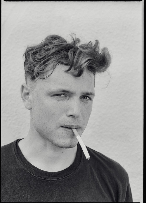 Monochrome portrait of a young adult man with styled hair, smoking a cigarette against a plain white background in a close-up shot.