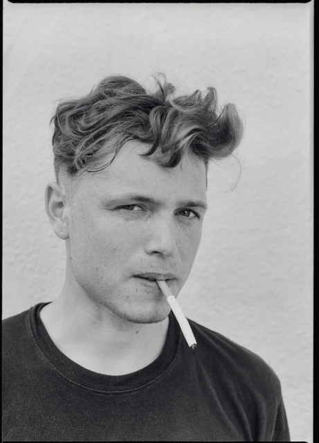 Monochrome portrait of a young adult man with styled hair, smoking a cigarette against a plain white background in a close-up shot.