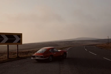 A vintage red sports car parked on the shoulder of a winding asphalt road beneath a hazy, overcast sky.