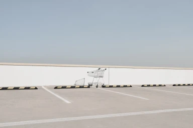 A wire shopping cart parked on an open concrete rooftop car park under a clear sky, photographed by Adam Kola.