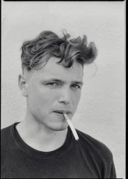 Portrait of a man with wavy hair and a cigarette in his mouth wearing a dark t-shirt against a textured white wall.