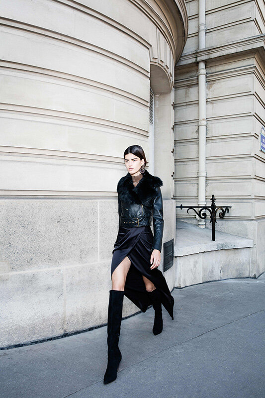 A stylish woman with a fur-collared leather jacket and black skirt walks along a city street with a stone wall background.