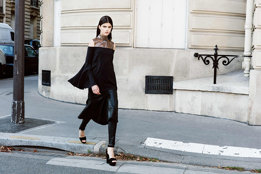 A woman in a black dress and leather pants walks on a city street near a building on a sunny day, showcasing a stylish fashion look.