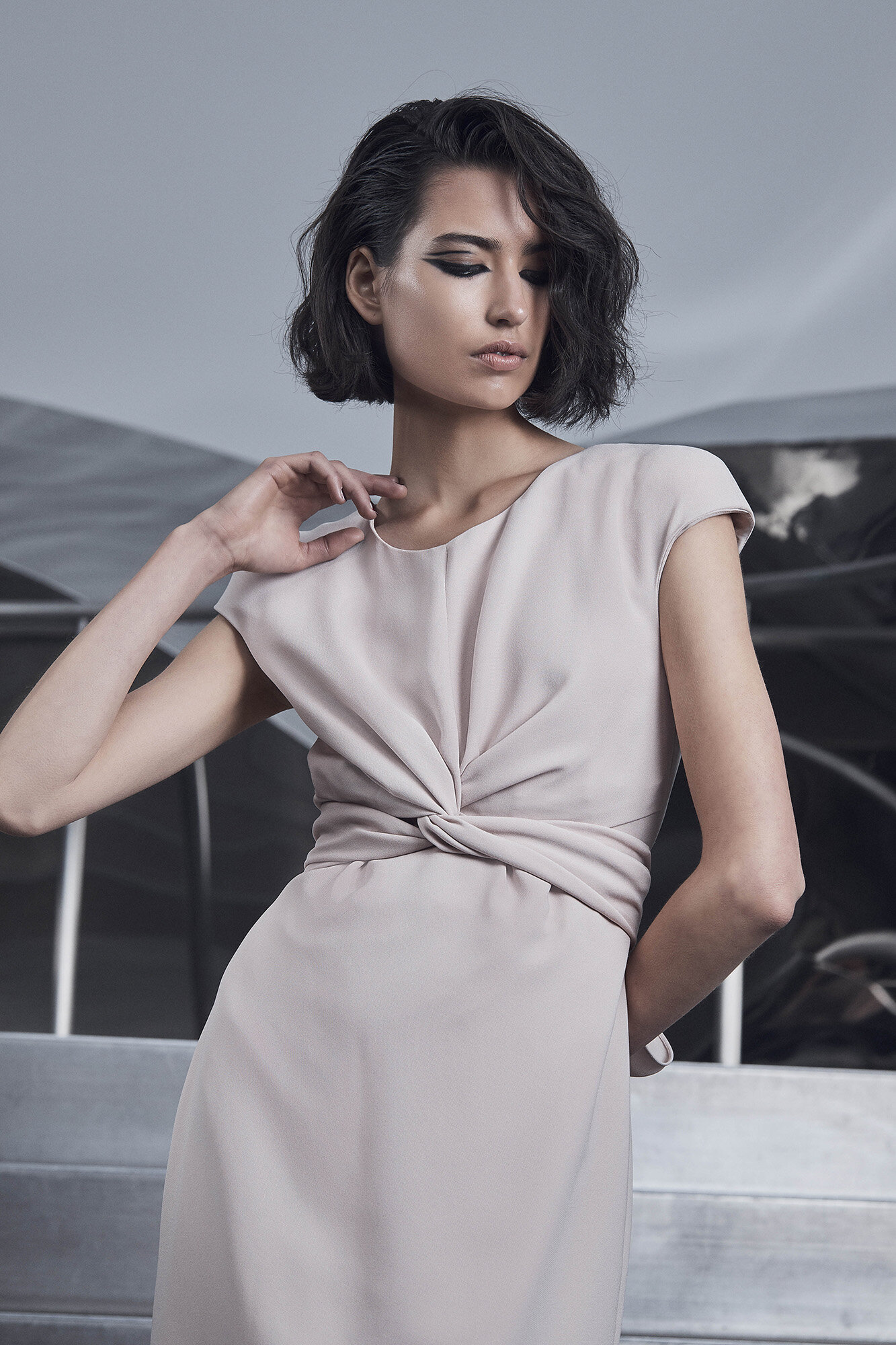 Elegant woman with bob hairstyle poses in a draped pale-pink dress against a geometric reflective backdrop in a fashion studio setting.