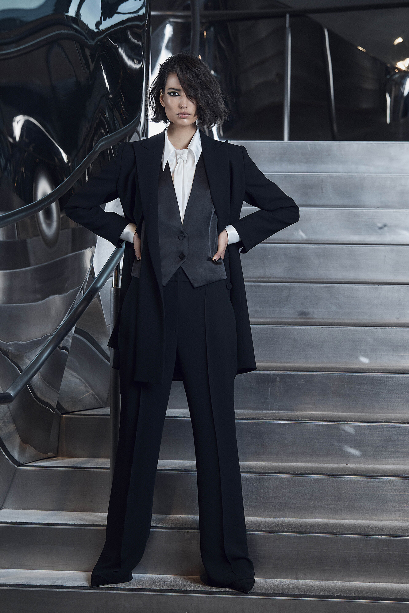 Woman in a stylish black suit with hands on hips posing on stairs with a metallic reflective background, showcasing fashion and elegance.