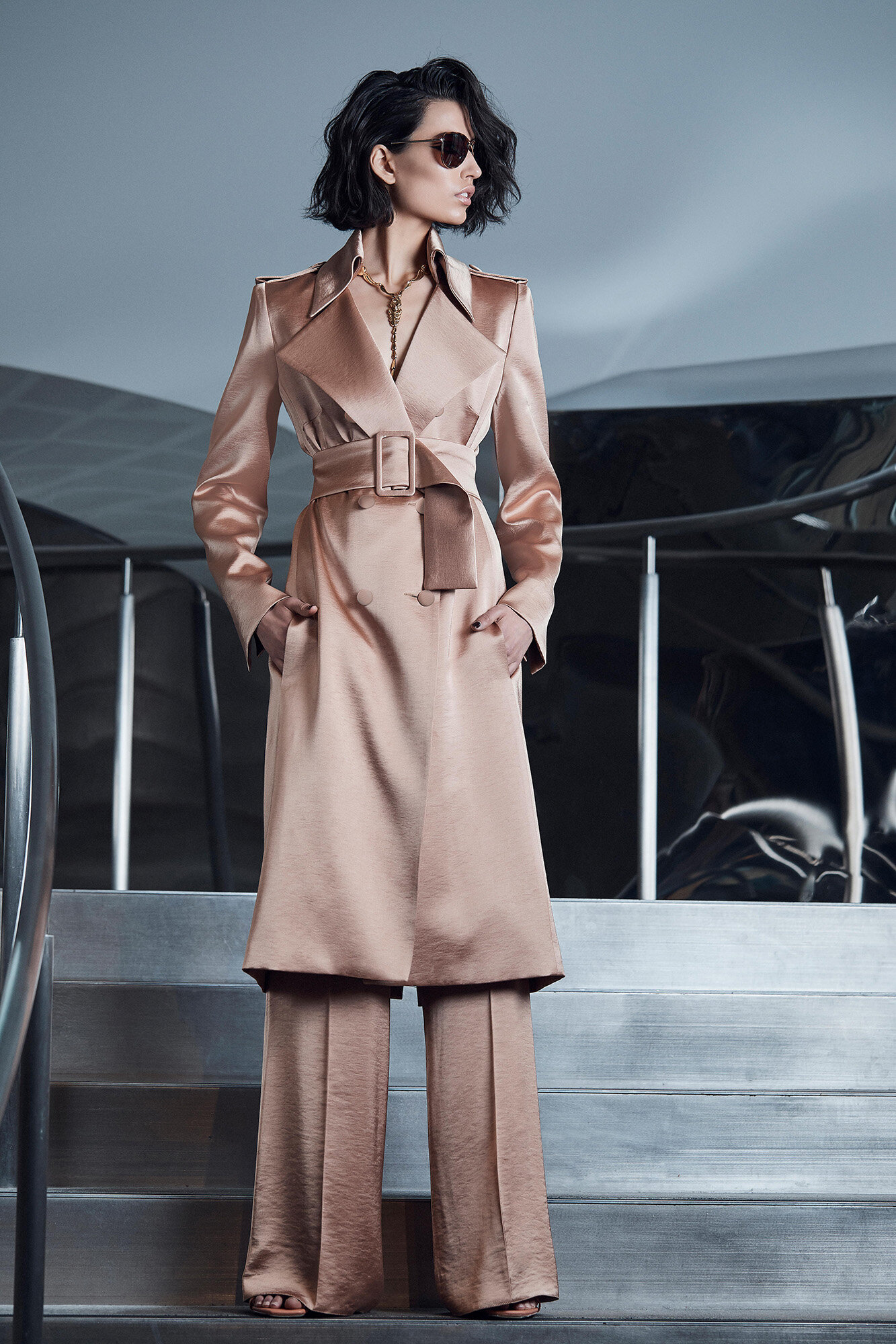Brunette model in sunglasses poses in a champagne-colored coat and matching pants on metal steps in a studio setting.