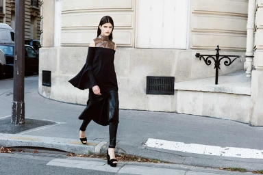 Model with long dark hair walking on a city street wearing an off-the-shoulder black dress and dark trousers.