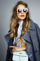Model wearing oversized geometric sunglasses, denim coat, floral blouse, light blue neck scarf, and holding a light blue handbag.