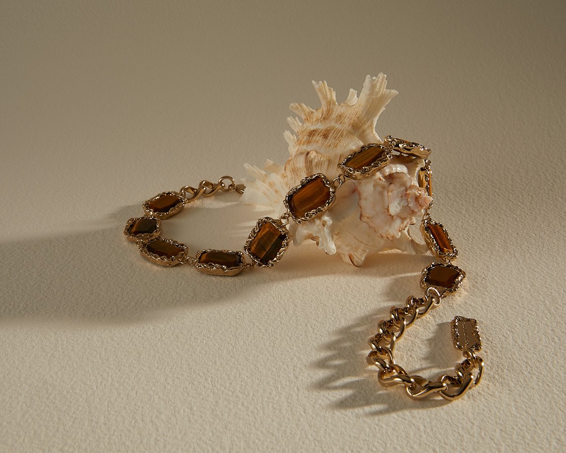 Golden chain bracelet with brown gemstones displayed next to a textured seashell on a light beige surface casting soft shadows.
