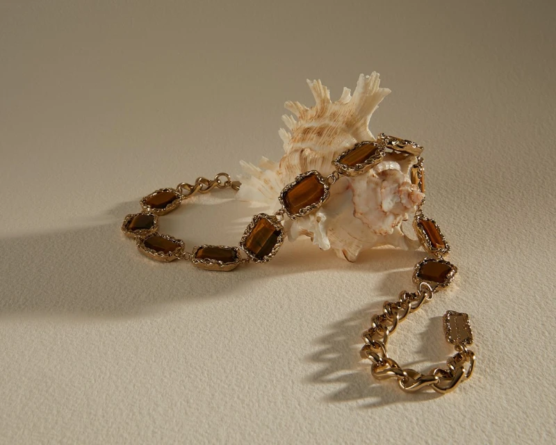 Golden chain bracelet with brown gemstones displayed next to a textured seashell on a light beige surface casting soft shadows.