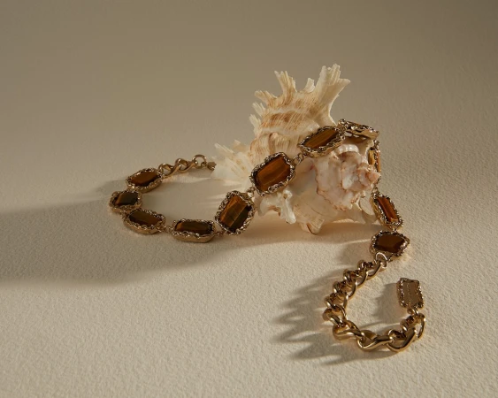 Golden chain bracelet with brown gemstones displayed next to a textured seashell on a light beige surface casting soft shadows.