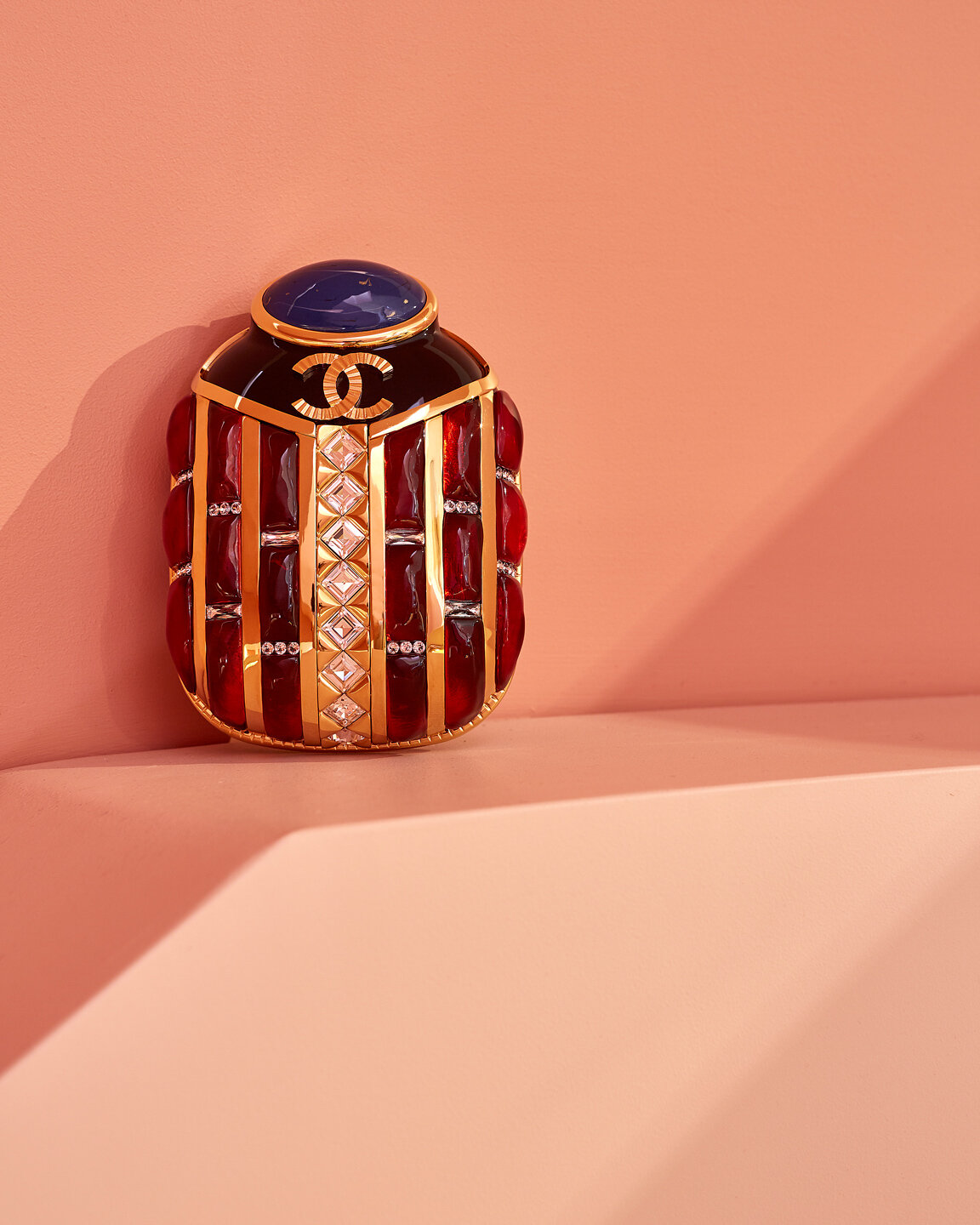 Still life of a decorative perfume bottle featuring red and gold details and a Chanel logo, set against a peach-colored background with a shadow.