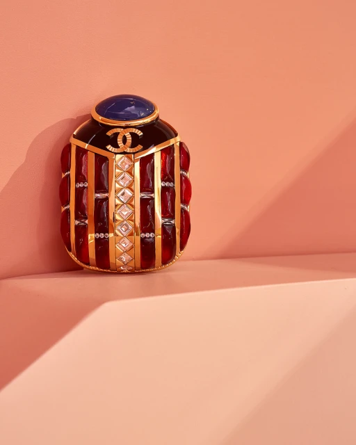 Still life of a decorative perfume bottle featuring red and gold details and a Chanel logo, set against a peach-colored background with a shadow.