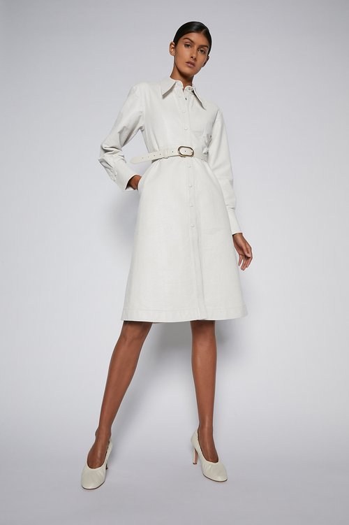 An adult female model poses in a cream-colored, long-sleeved dress with a belt and matching high heels on a bright white background.