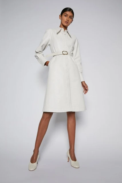 An adult female model poses in a cream-colored, long-sleeved dress with a belt and matching high heels on a bright white background.