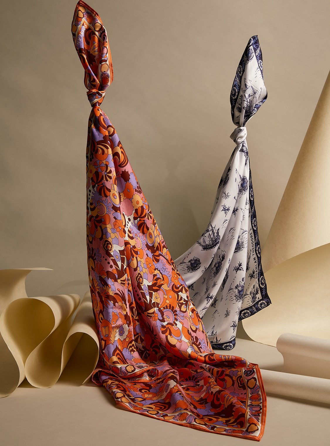 Two patterned silk scarves knotted at the top, draped in soft folds with a minimalist paper-art background in a fashion still life.