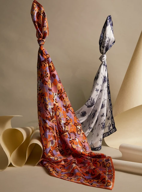 Two patterned silk scarves knotted at the top, draped in soft folds with a minimalist paper-art background in a fashion still life.