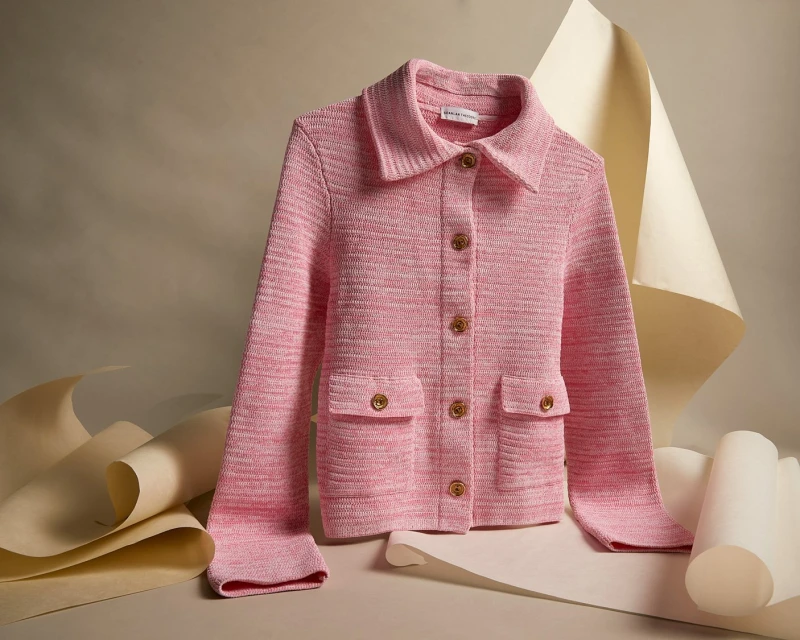 Pink knit jacket with wood buttons and front pockets against a neutral studio backdrop with curled beige paper accents.