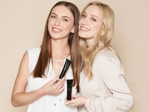 Two smiling women pose in a studio with cosmetic product tubes. Beauty shot for skincare advertising with neutral background and soft lighting.