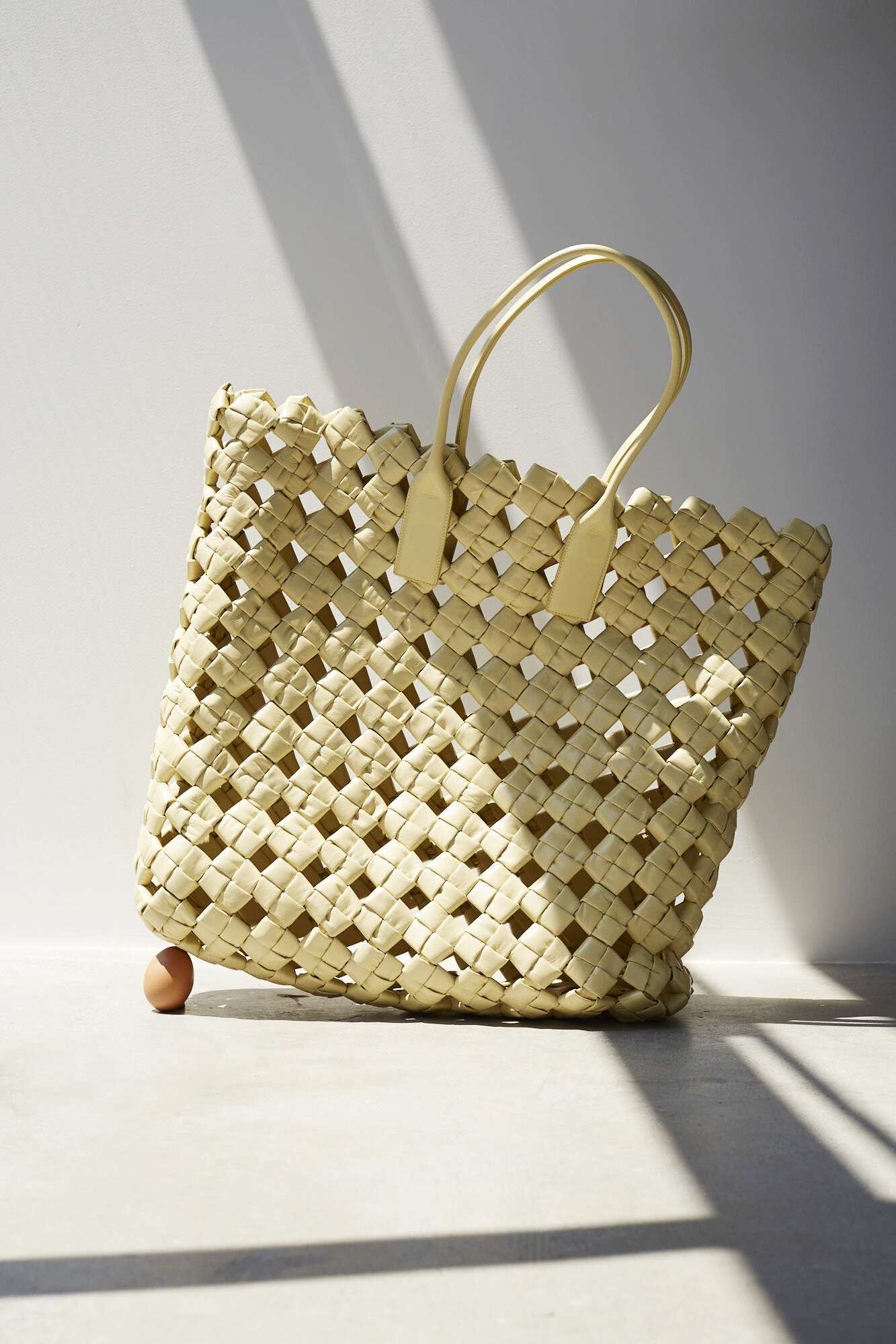 Woven straw tote bag with brown leather handles and a single brown egg on a light grey background with shadows.