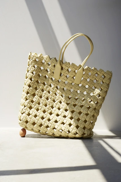 Woven straw tote bag with brown leather handles and a single brown egg on a light grey background with shadows.
