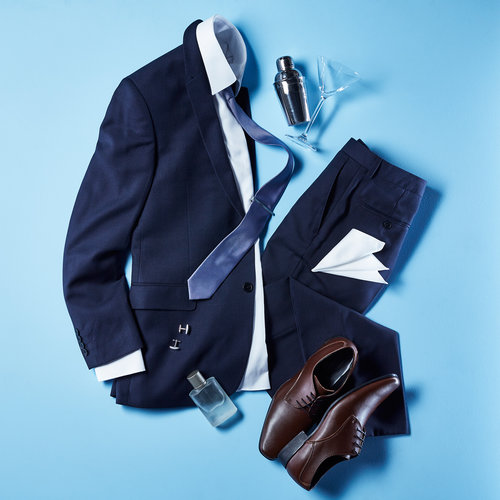 Arrangement of a men's navy suit, dress shirt, tie, leather shoes, and accessories, with a cocktail shaker on a light blue background.
