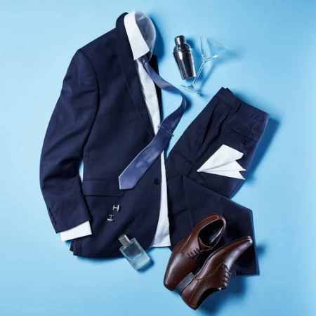 Arrangement of a men's navy suit, dress shirt, tie, leather shoes, and accessories, with a cocktail shaker on a light blue background.
